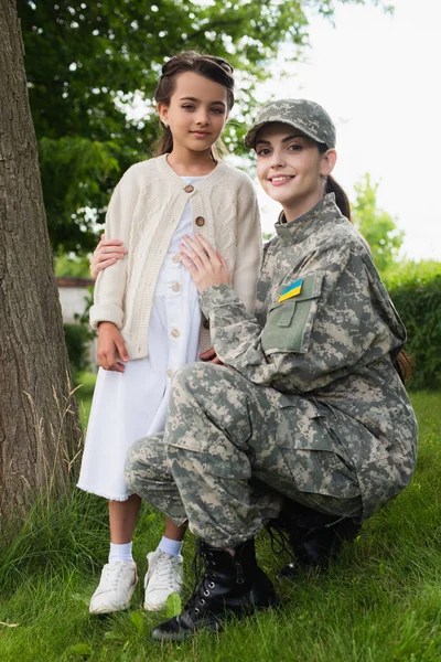 Happy woman in camouflage embracing daughter and looking at camera in park — стоковое фото