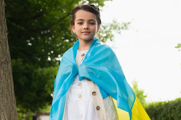 Girl covered with ukrainian flag looking at camera outdoors — Photo de stock