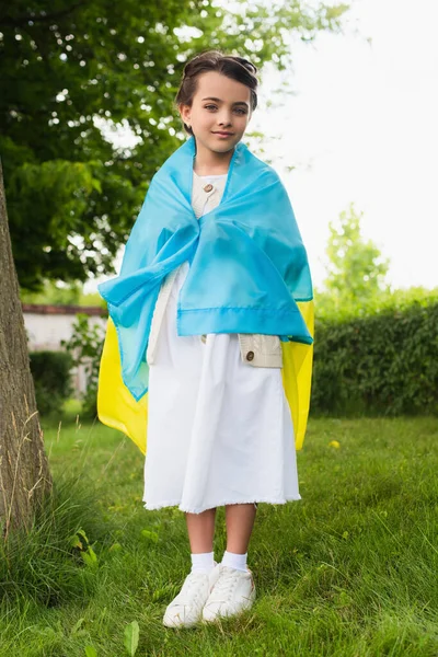 Full length of patriotic child covered with ukrainian flag in park — стоковое фото