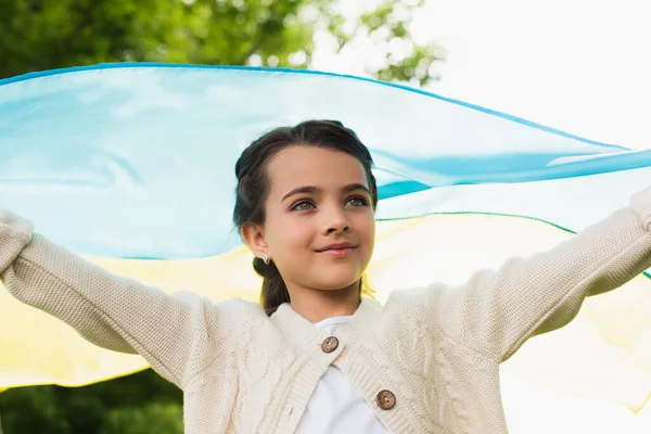 Positive patriotic girl with ukrainian flag looking away outdoors — стоковое фото