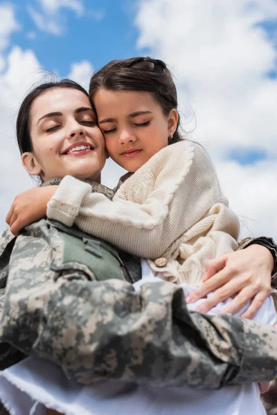 Smiling military woman with closed eyes embracing daughter against cloudy sky — Photo de stock