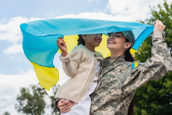 Усміхнена жінка в камуфляжі тримає український прапор з дочкою на відкритому повітрі — стокове фото