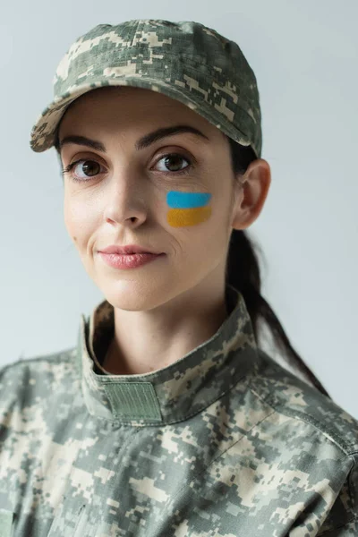 Military woman with ukrainian flag painted on face looking at camera isolated on grey — Photo de stock