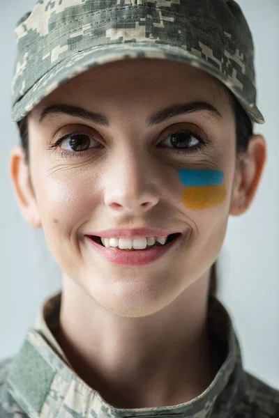 Close up portrait of military woman with ukrainian flag on face smiling at camera isolated on grey - foto de stock