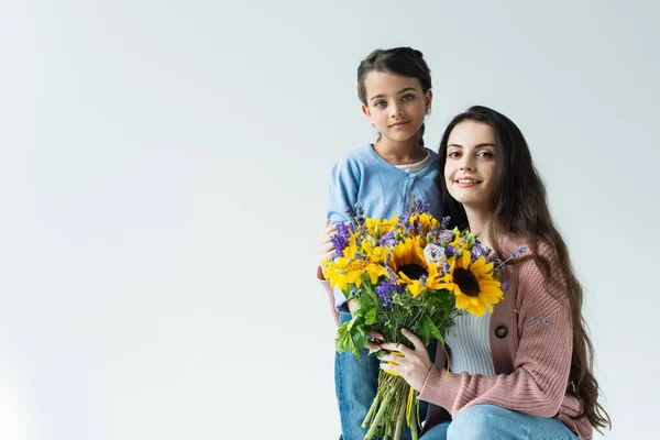 Жінка і дівчина посміхаються на камеру біля букета синьо-жовтих квітів ізольовані на сірому — стокове фото