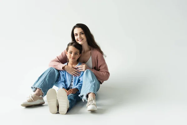 Full length of cheerful woman hugging daughter while sitting on grey background - foto de stock