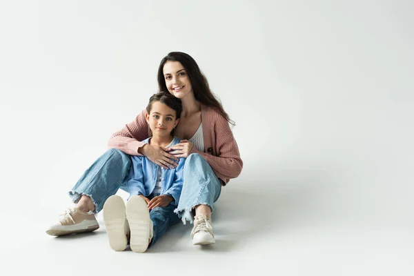 Full length of cheerful woman hugging daughter while sitting on grey background — стоковое фото