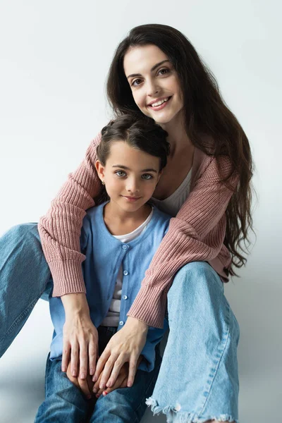 Joyful brunette woman embracing daughter and looking at camera on grey background — Stock Photo