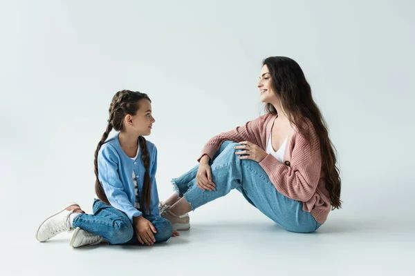 Happy woman looking at daughter with pigtails sitting on grey background — стоковое фото