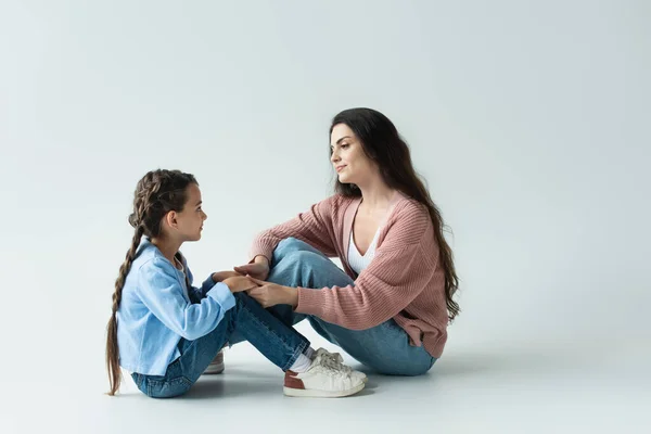 Side view of mom and daughter in jeans holding hands while sitting on grey background — стоковое фото
