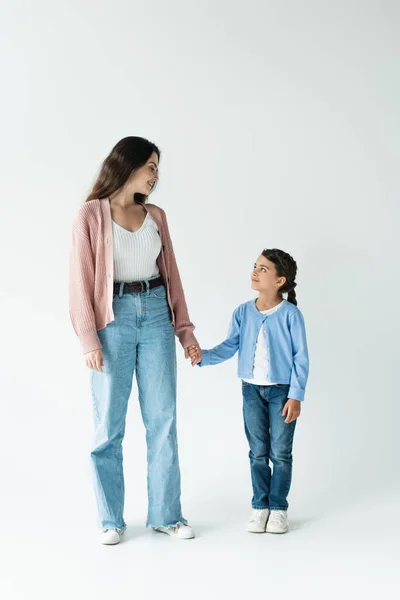 Full length of woman and girl in jeans holding hands and smiling at each other on grey background - foto de stock