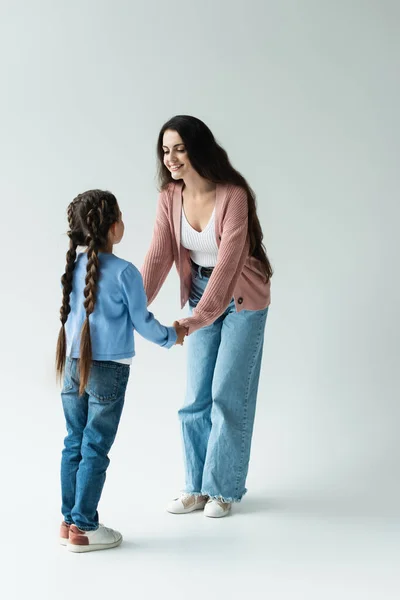 Full length of cheerful woman holding hands of daughter with pigtails on grey background — стоковое фото