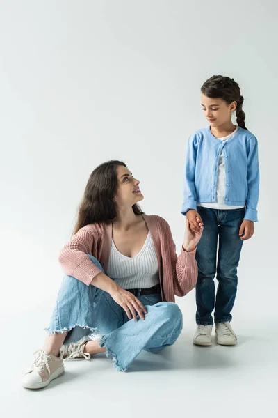 Brunette woman in jeans holding hand of daughter while sitting on grey background — Photo de stock