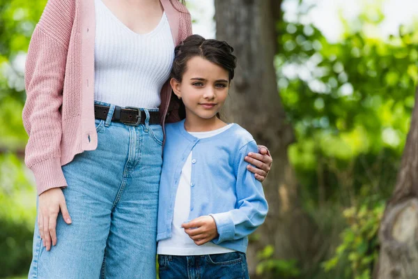 Girl looking at camera near mother hugging her outdoors — стоковое фото