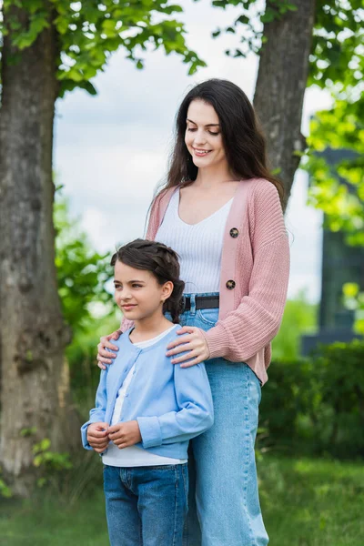 Smiling woman embracing shoulders of pensive daughter in park — стоковое фото