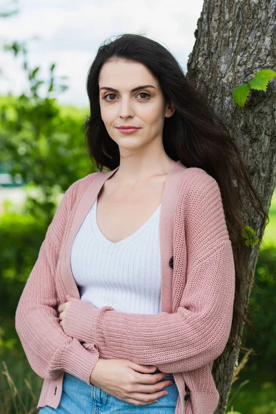 Positive brunette woman standing with crossed arms in park and looking at camera — стоковое фото