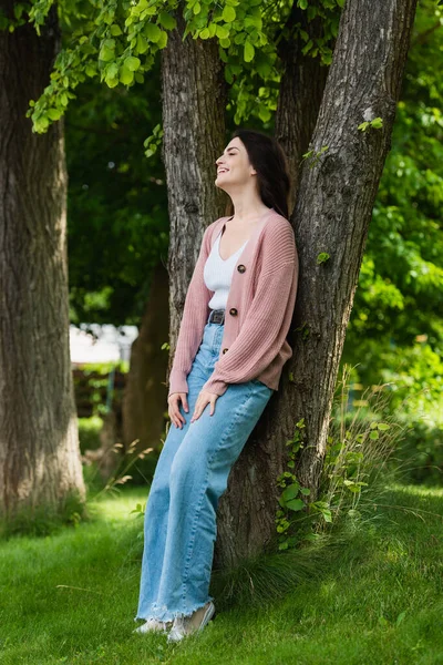 Brunette woman in jeans smiling near trees in park — Stockfoto