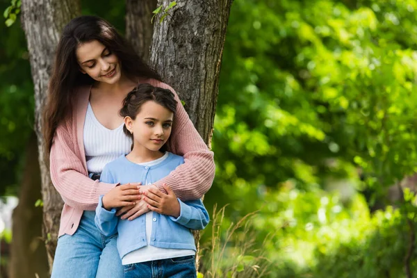 Щаслива молода жінка обіймає дочку біля дерев у парку — стокове фото