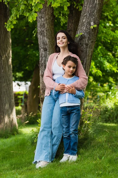 Full length of positive woman hugging daughter near trees in park — Foto stock