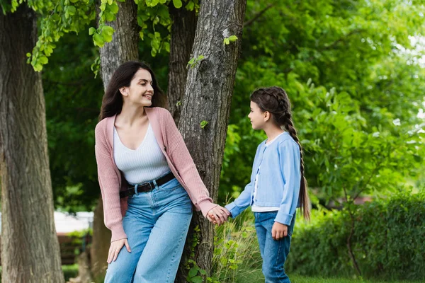 Young woman and child holding hands and looking at each other near trees in park — Fotografia de Stock