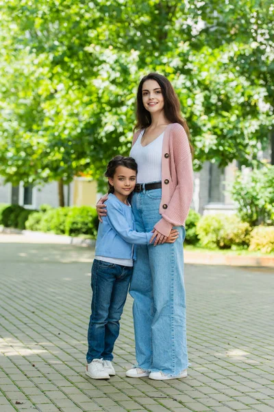 Full length of mom and daughter in jeans embracing and smiling at camera on city street — Stockfoto