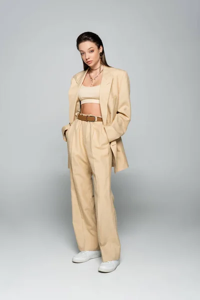 Full length of pretty young woman in beige blazer posing with hands in pockets on grey — Photo de stock