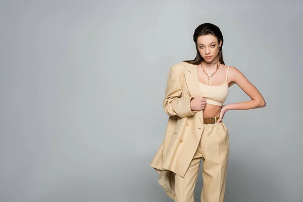 Pretty young woman in beige blazer looking at camera while posing with hand on hip isolated on grey - foto de stock