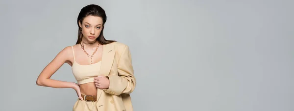 Pretty young woman in beige blazer looking at camera while posing with hand on hip isolated on grey, banner - foto de stock