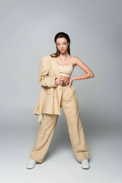 Full length of stylish young woman in beige blazer looking at camera while posing with hand on hip on grey — стоковое фото