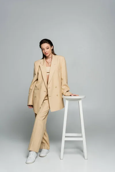 Full length of pretty woman in beige blazer looking at camera while posing near high stool on grey — Photo de stock