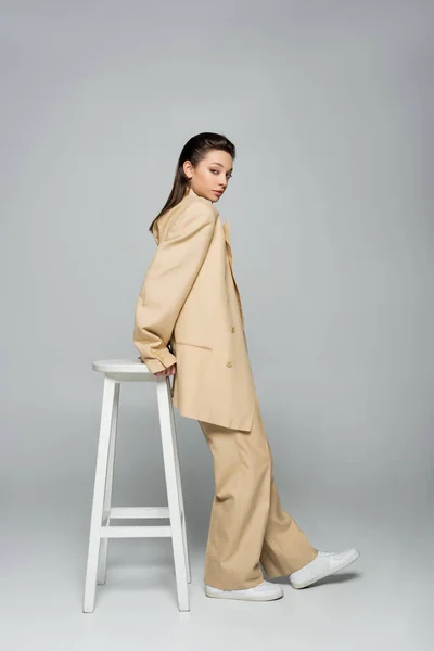 Full length of young woman in beige suit looking at camera while posing near high stool on grey — Fotografia de Stock