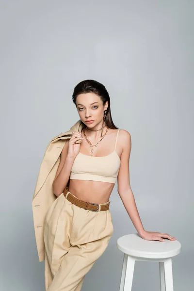 Young woman in beige outfit holding blazer and looking at camera while posing near high stool isolated on grey — стоковое фото