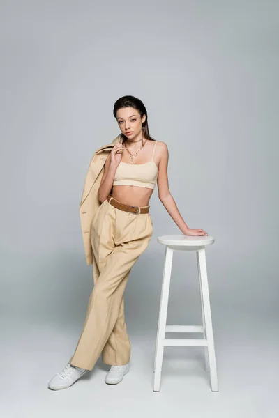Full length of young woman in beige outfit holding blazer and posing near high stool on grey - foto de stock