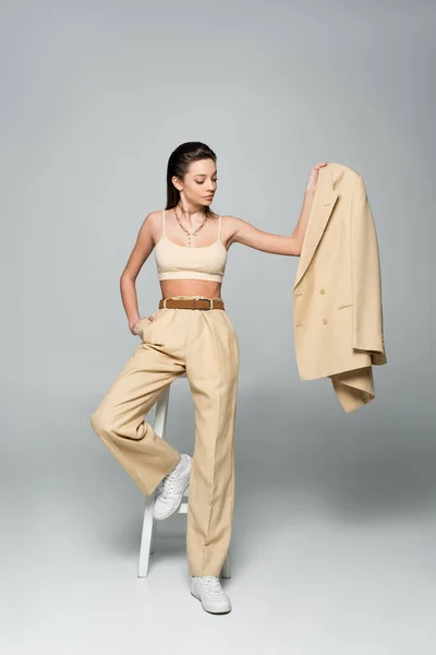 Full length of brunette model in beige outfit holding blazer and posing with hand in pocket near high stool on grey — Fotografia de Stock