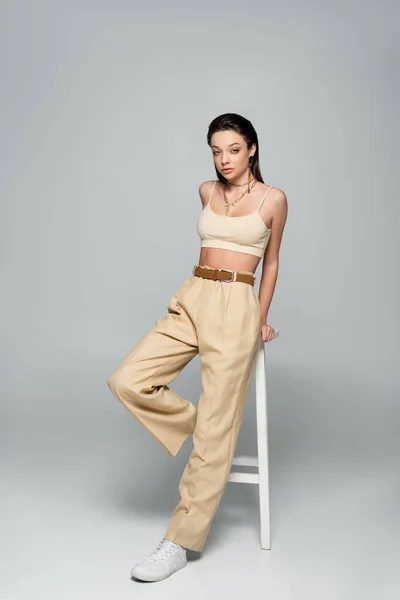 Full length of pretty young woman in beige outfit looking at camera while posing near high stool on grey — Stock Photo