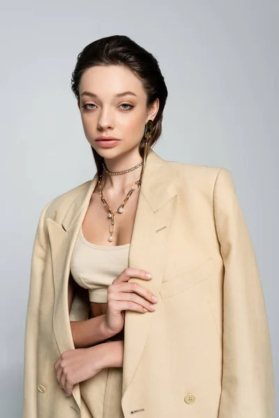 Young woman in beige outfit holding jacket and looking at camera while posing isolated on grey — Stock Photo