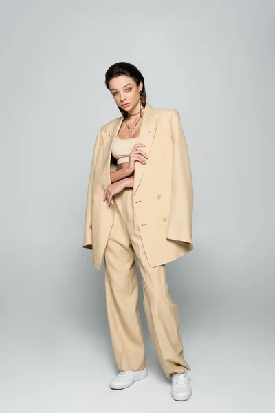 Full length of pretty young woman in beige outfit looking at camera while posing on grey — Stockfoto