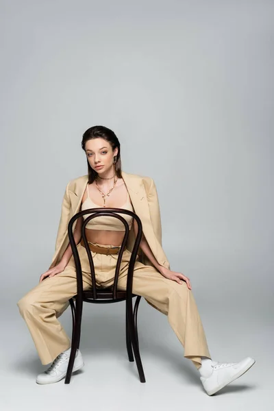 Full length of young woman in beige outfit looking at camera while sitting on wooden chair on grey — Foto stock