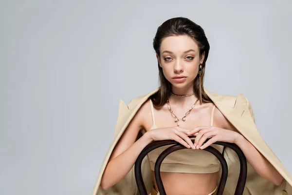 Young woman in beige blazer looking at camera and sitting on wooden chair isolated on grey — Stockfoto