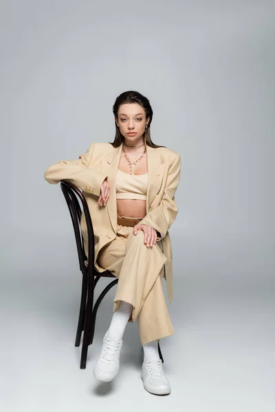 Full length of young woman in stylish outfit looking at camera while posing on wooden chair on grey — Stockfoto