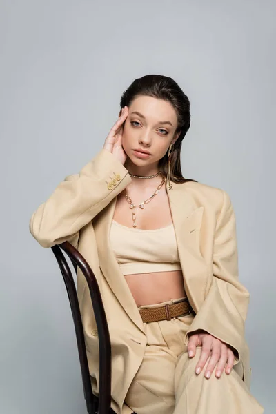 Trendy woman in stylish outfit looking at camera while sitting on chair isolated on grey — Fotografia de Stock