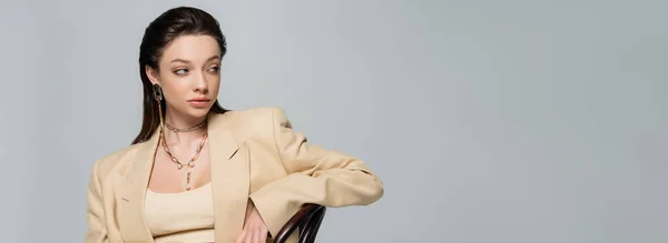 Brunette young woman in stylish outfit looking away and leaning on chair isolated on grey, banner — стоковое фото