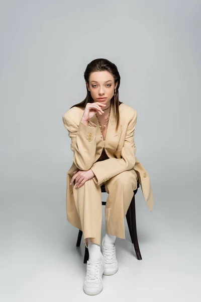 Full length of stylish woman in trendy outfit looking at camera while sitting on chair on grey - foto de stock