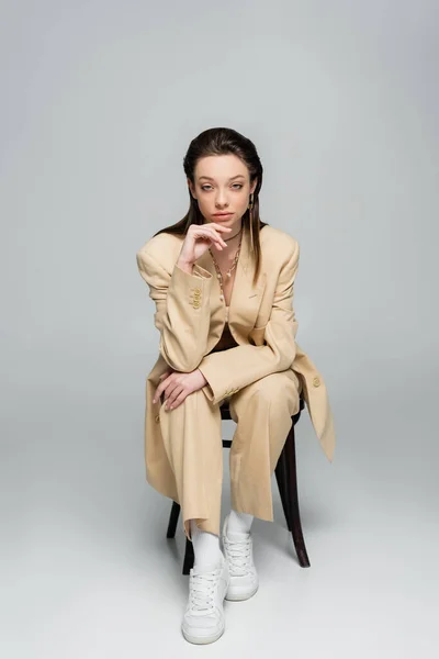 Full length of stylish woman in trendy outfit looking at camera while sitting on chair on grey — стоковое фото