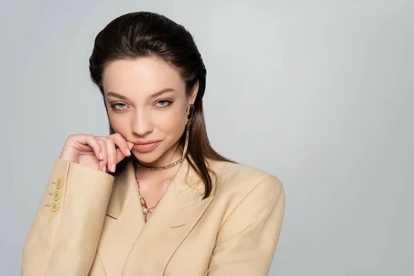 Pleased young woman in stylish blazer looking at camera while posing isolated on grey — стоковое фото