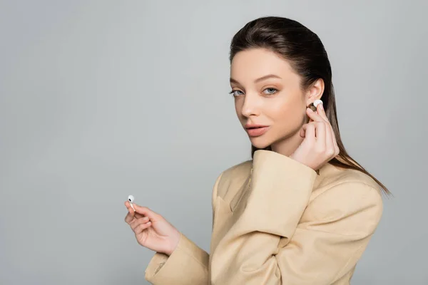 Young woman in stylish outfit looking at camera while wearing wireless earphones isolated on grey — стоковое фото