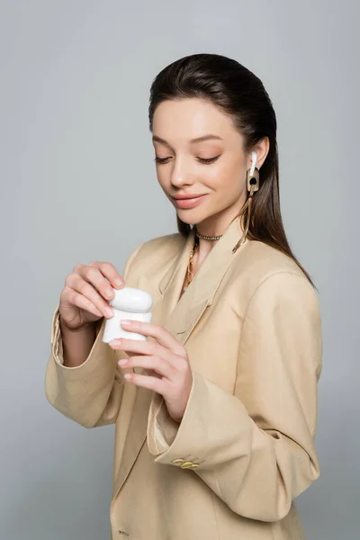 Happy woman in stylish outfit looking at plastic case while listening music in earphone isolated on grey — Stock Photo