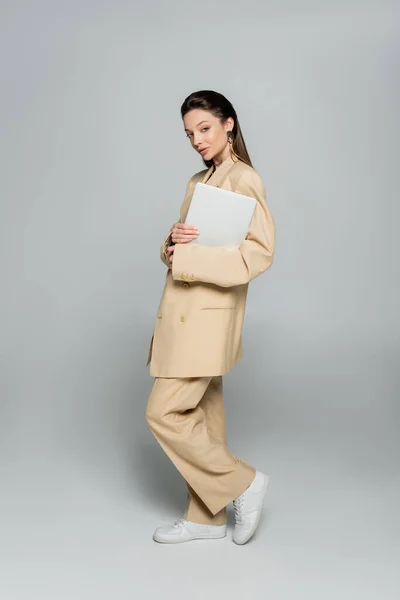 Full length of young woman in stylish outfit looking at camera while holding laptop on grey — Stock Photo