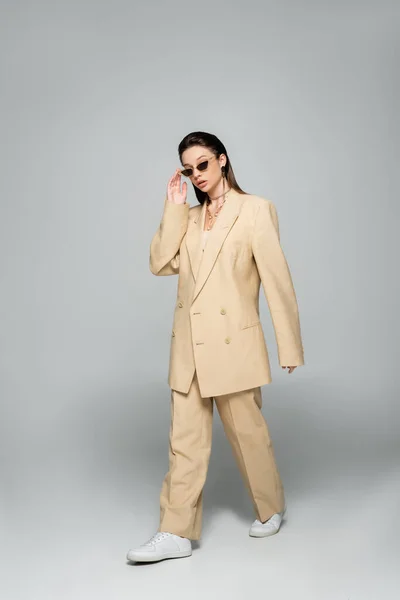Full length of young woman in stylish beige suit wearing trendy sunglasses and walking on grey — стоковое фото