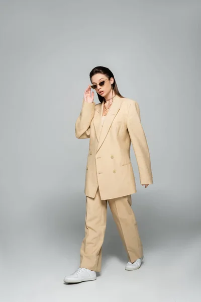 Full length of young woman in stylish beige suit wearing trendy sunglasses and walking on grey — Stock Photo